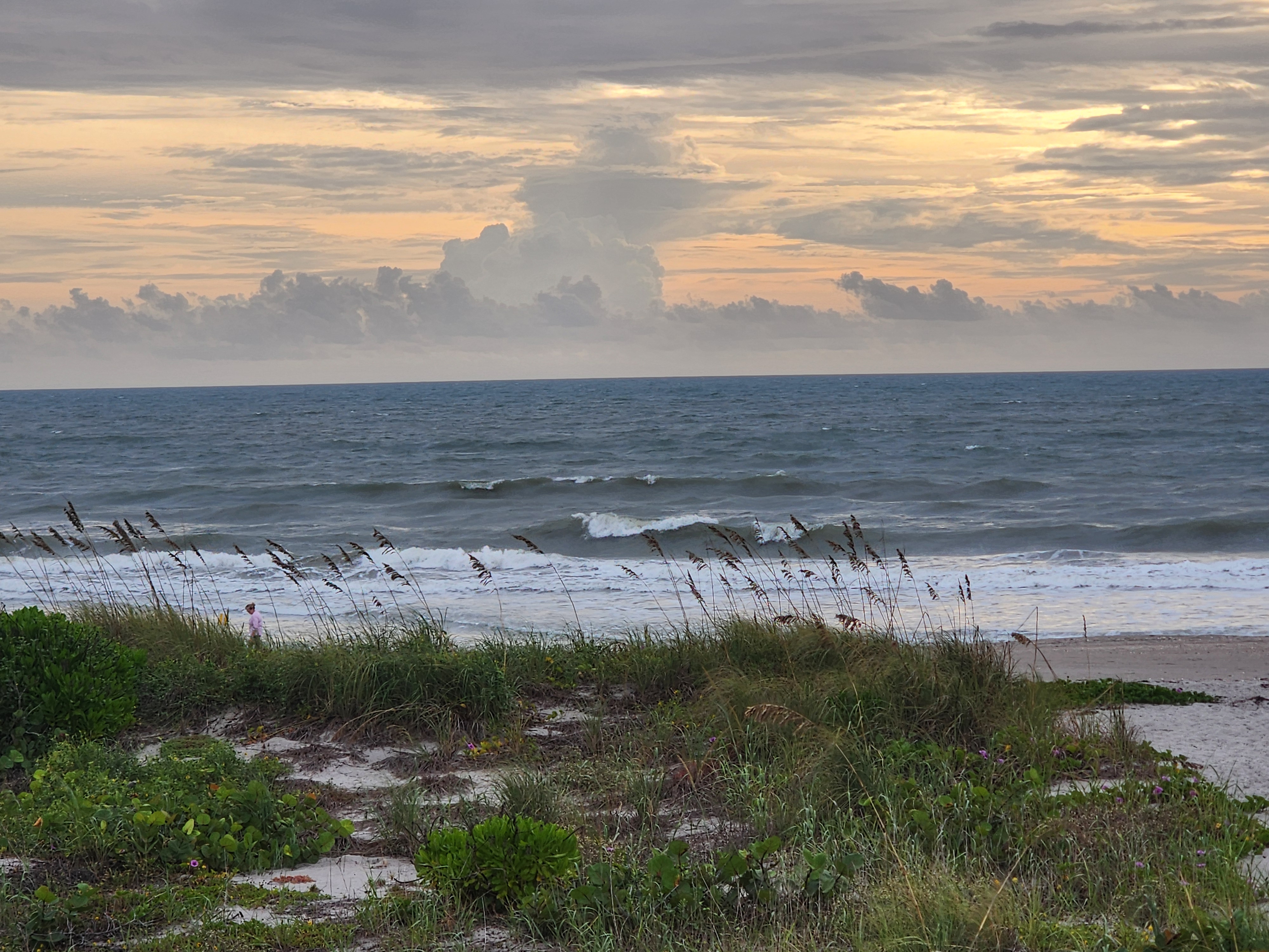 Peach and gray morning sky with a tall cumulus tower over the horizon, choppy waves breaking in several lines, and a lone beach walker in pink just past the dunes