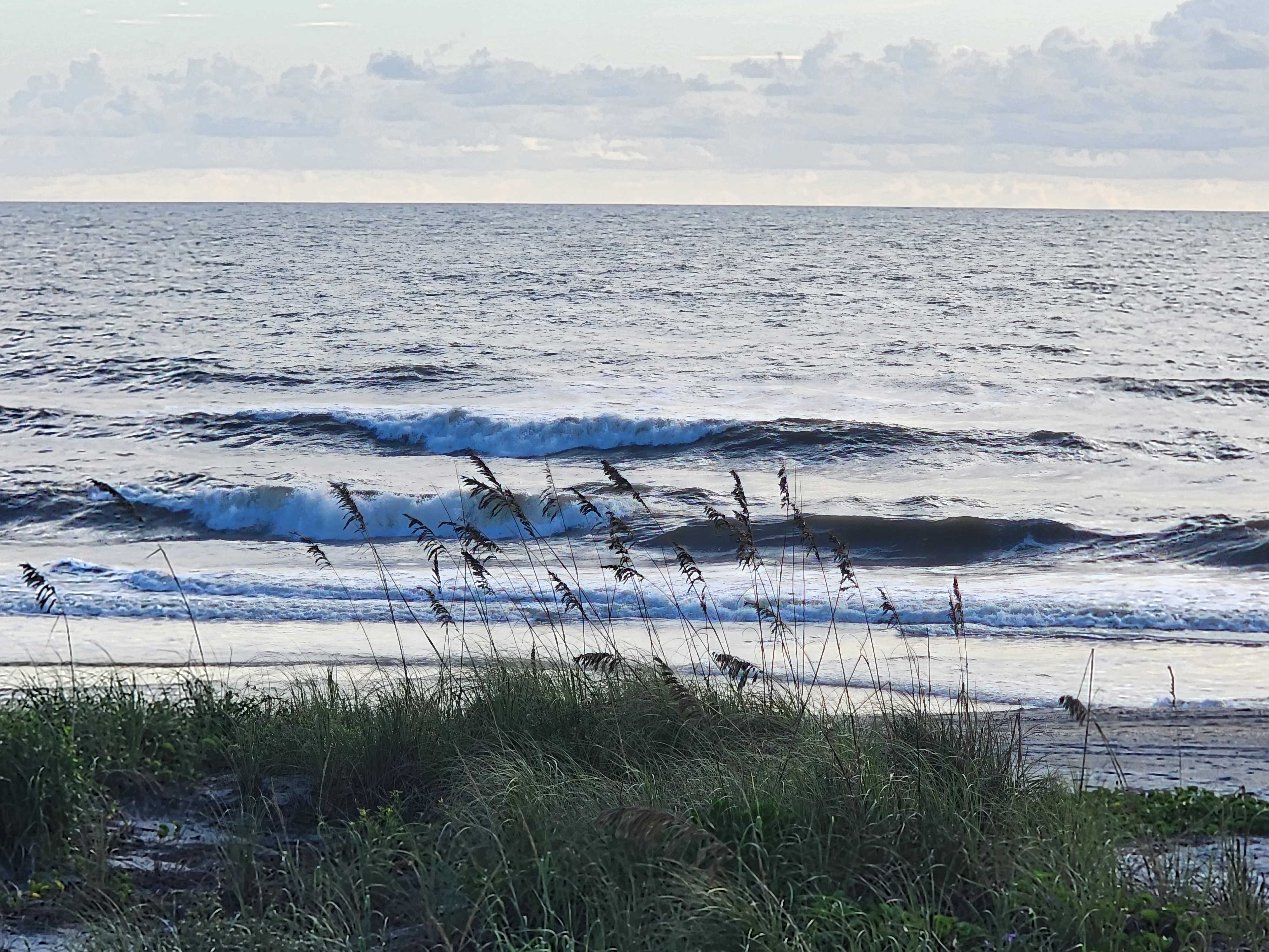 Two lines of 2-3 foot waves breaking over the shallow sandbar at Cocoa Beach, framed by silhouetted sea oats under a soft morning sky with scattered clouds