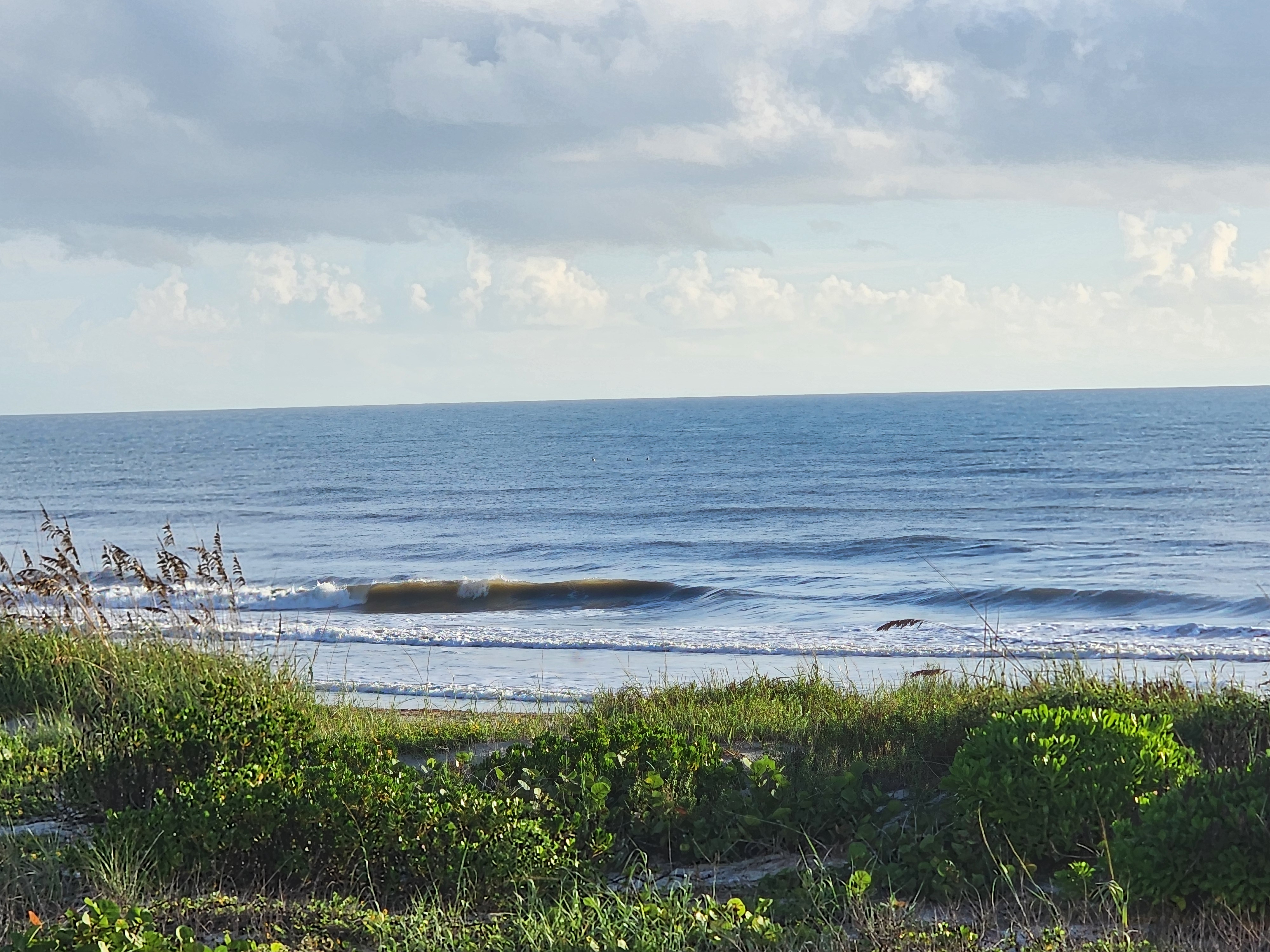 Lone longboard-sized wave standing up with a clean shoulder on the sandbar, light SW winds grooming the surface beneath scattered cumulus clouds