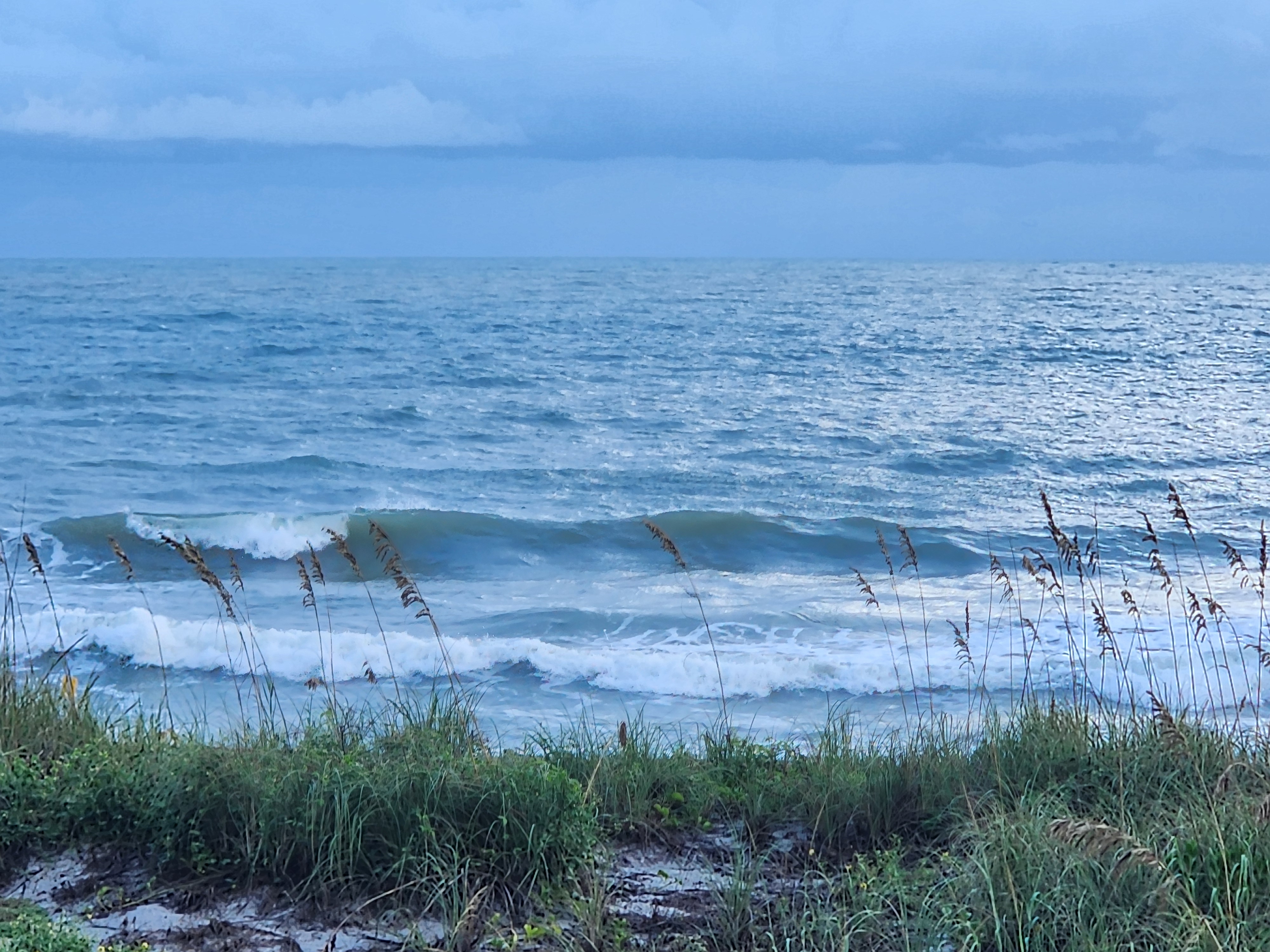 Two lines of chest-high surf breaking under a slate-blue sky, the outer wave pitching white on the left while sea oats lean across the foreground on the Cocoa Beach dune