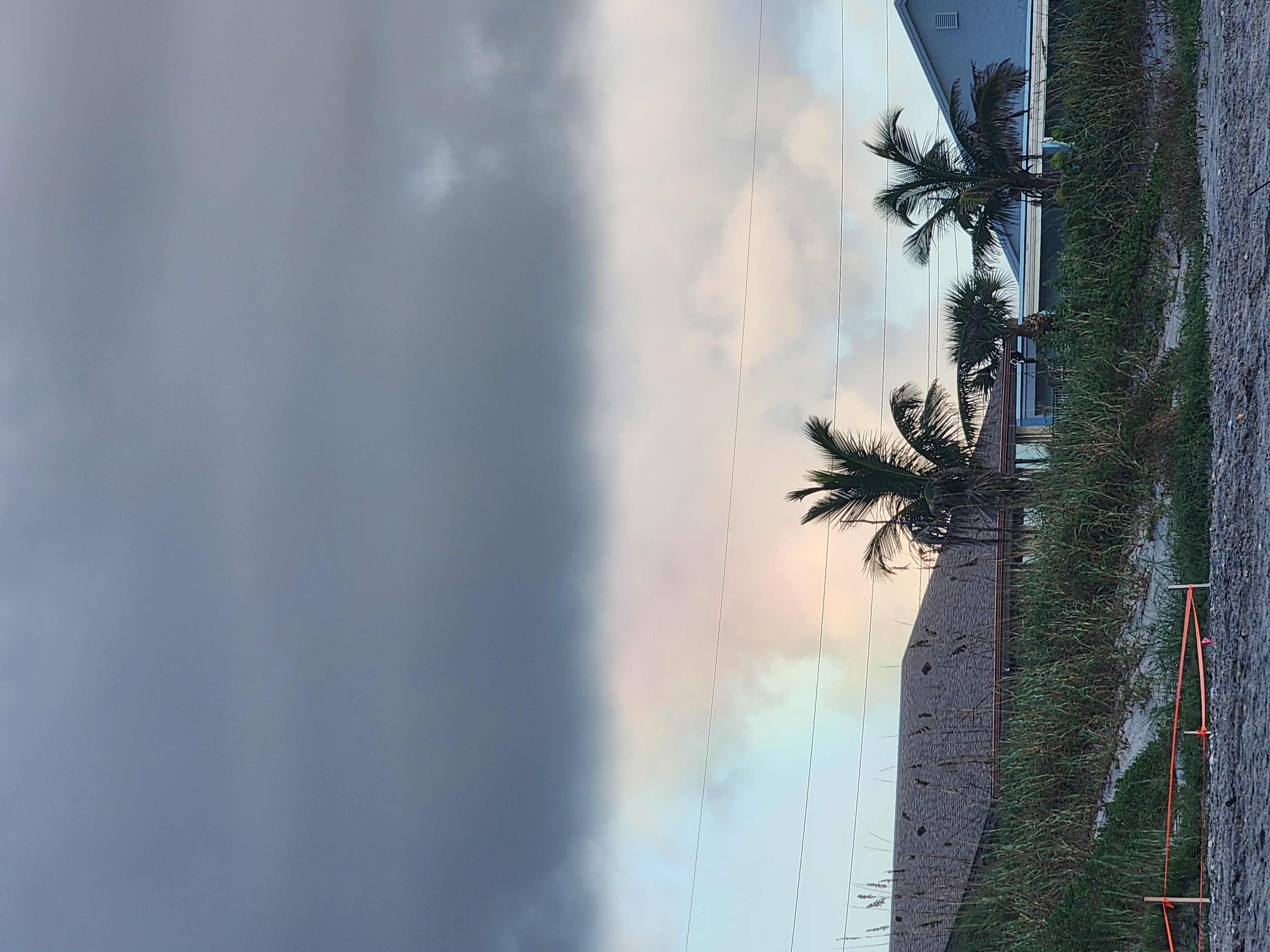 Faint rainbow glowing through gray clouds above palm trees and beachfront rooftops, orange-taped turtle nest stakes in the foreground
