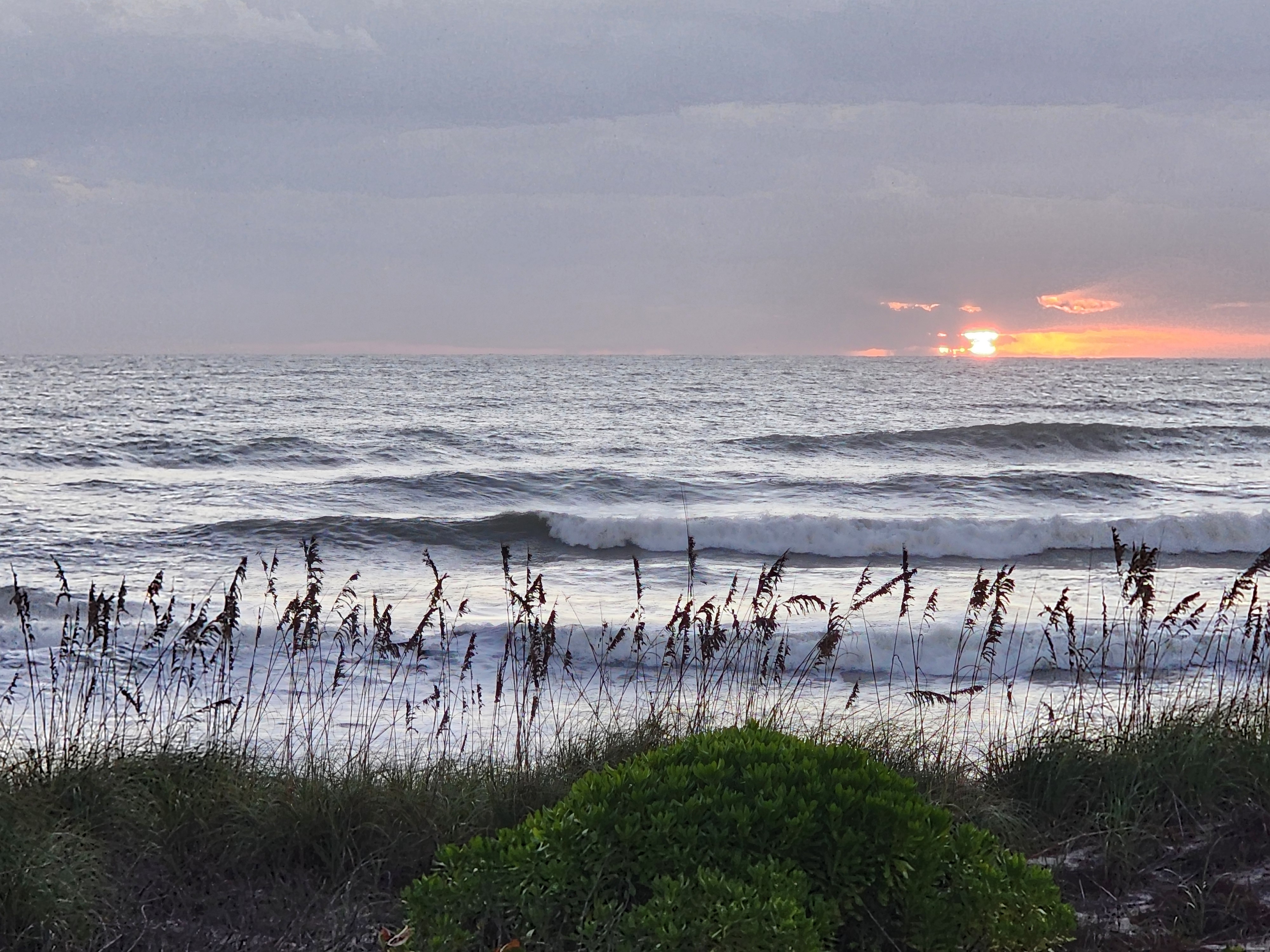 A blazing orange sunrise slipping out beneath a gray cloud deck on the right, silver light across a choppy ocean, and a long wave peeling into whitewater behind a fringe of backlit sea oats and a rounded green dune shrub