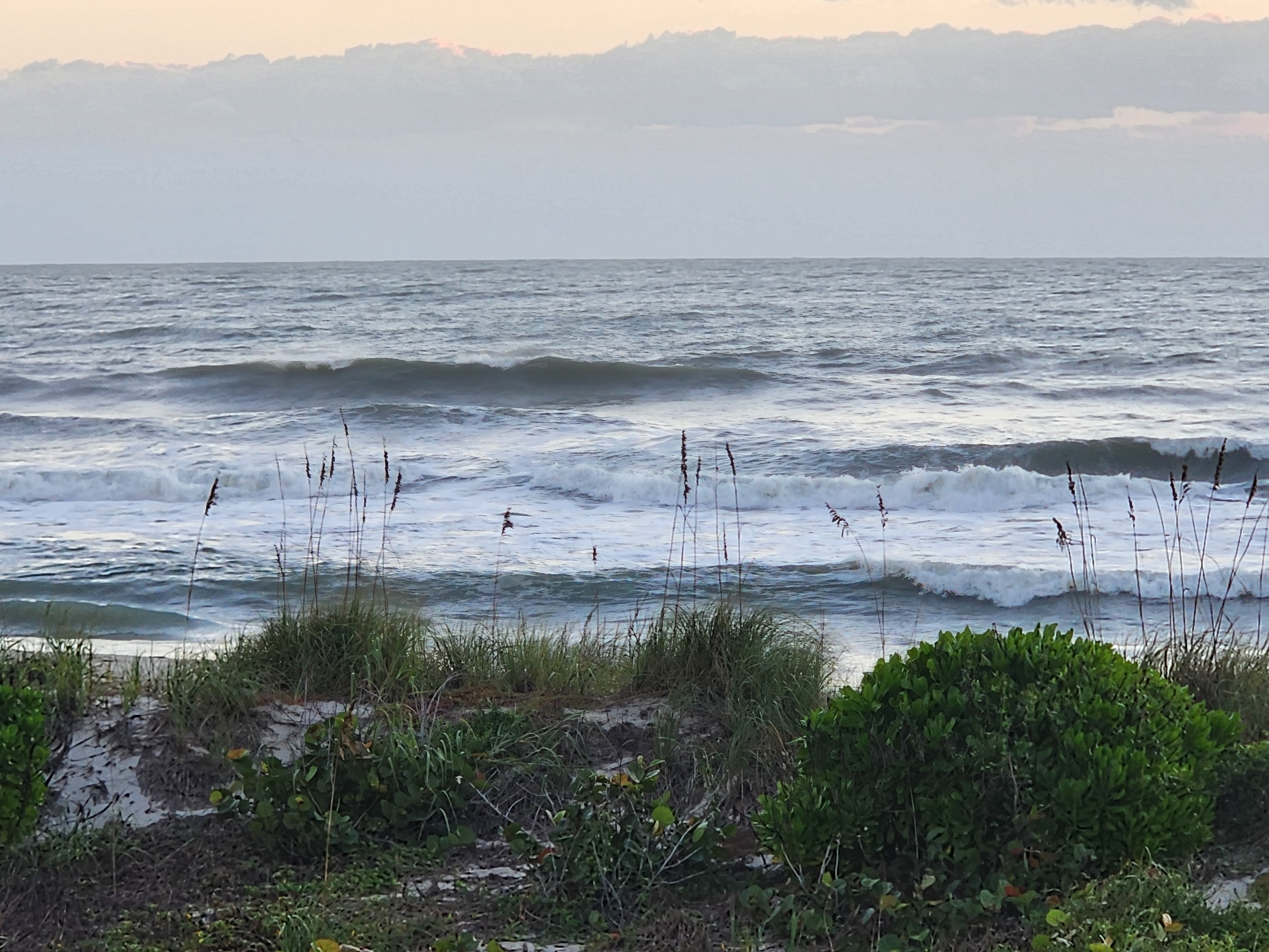 Several lines of jumbled surf stacking toward shore under a soft pastel sky, a clean lump standing up on the outside and whitewater churning across the inside, framed by sparse sea oats and dune shrubs