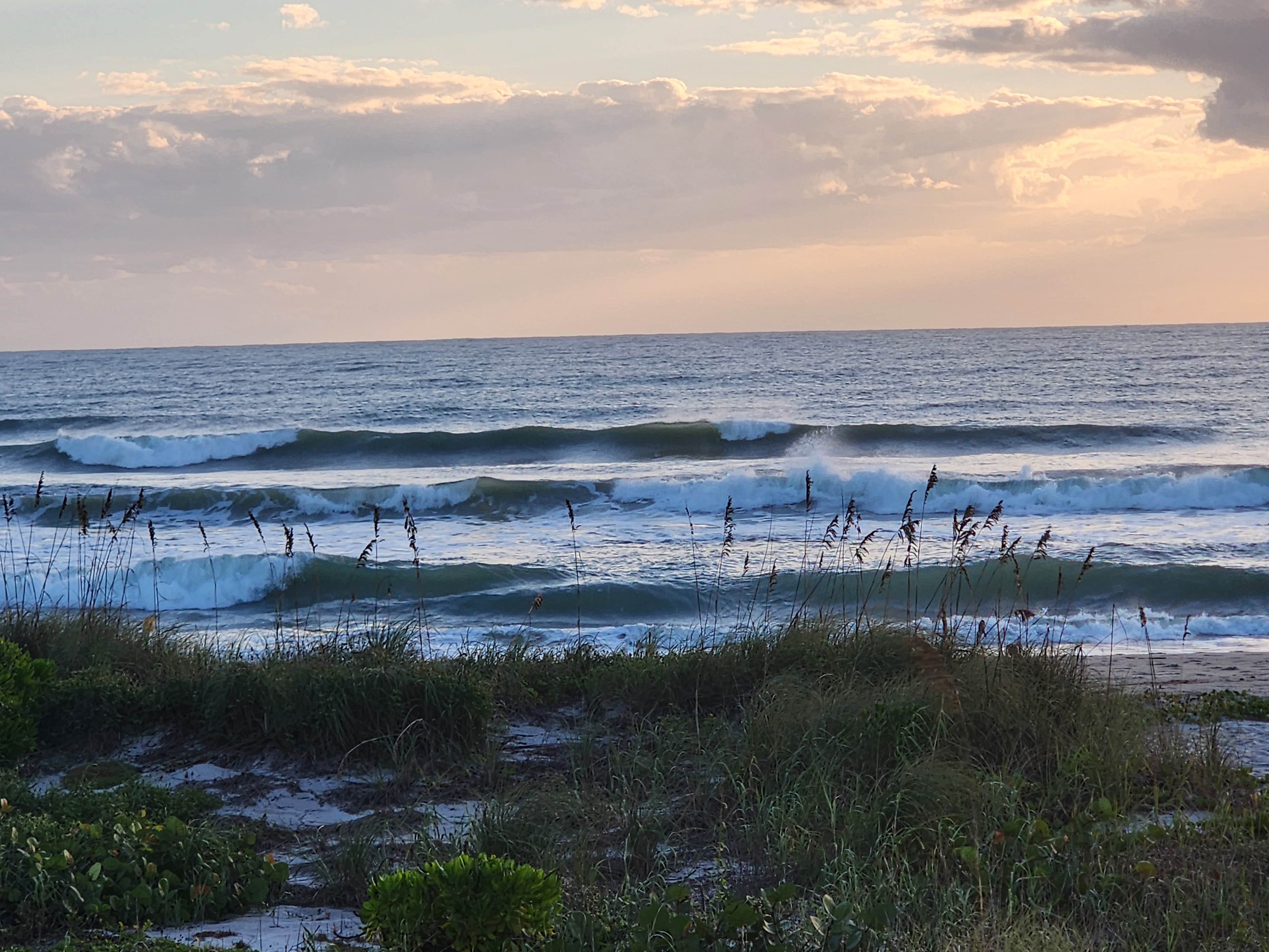 Three rows of breaking waves under peach-colored clouds, offshore winds blowing mist back off the peaks