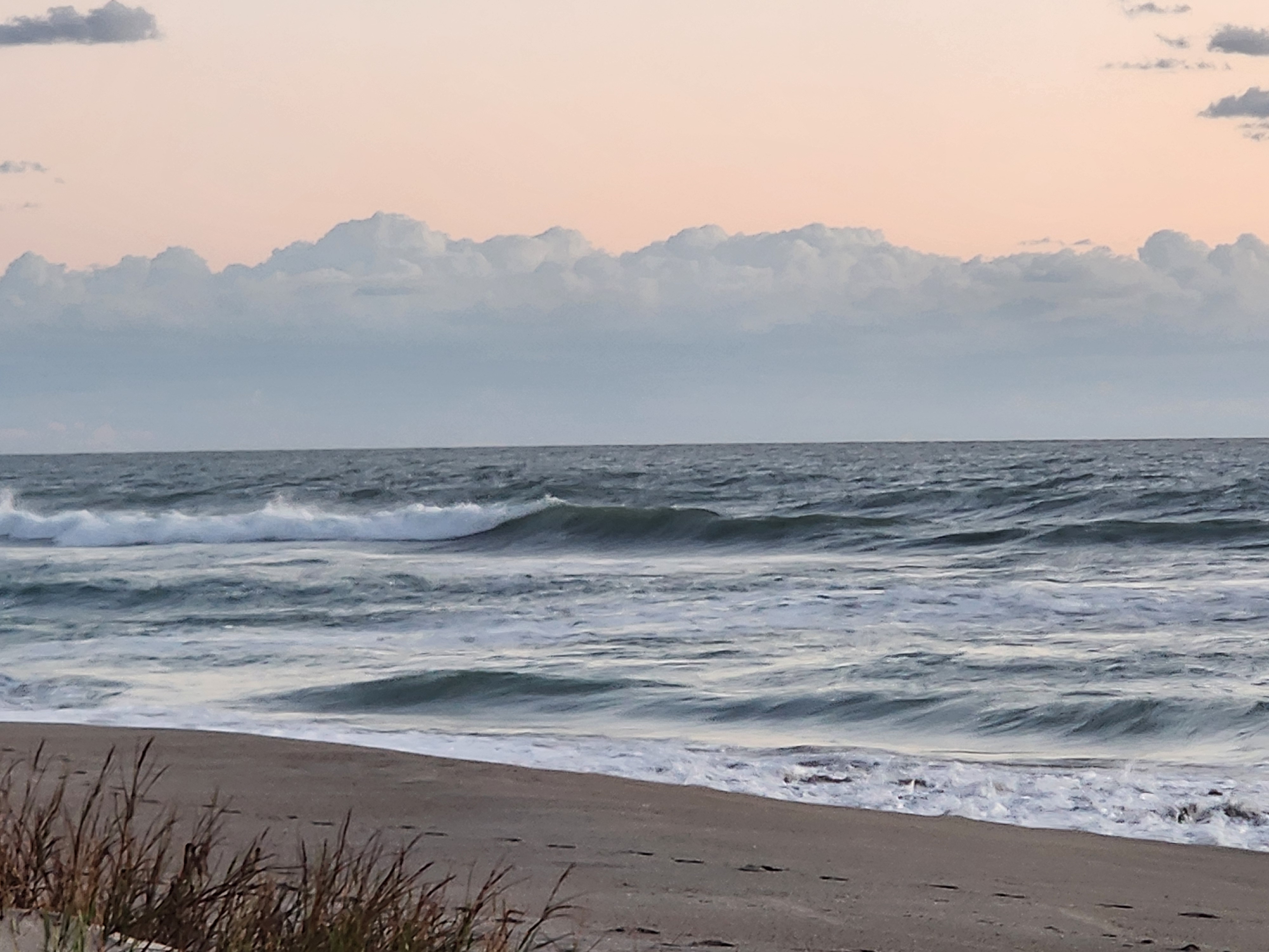 Shoulder-high waves breaking left across the outer bar with whitewater rolling toward the shore under pastel morning clouds
