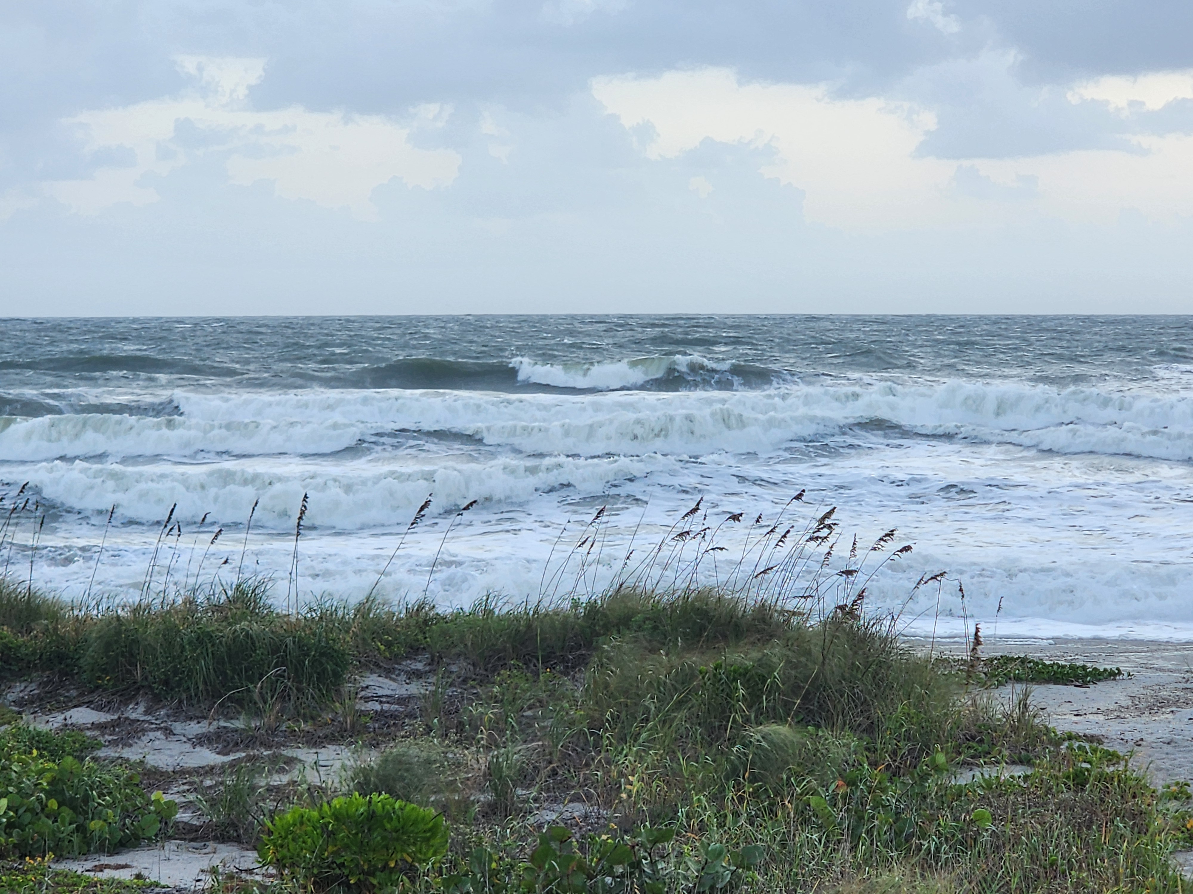 Stacked walls of whitewater and a peak pitching on the outer bar, king tide washing up near the dune grass